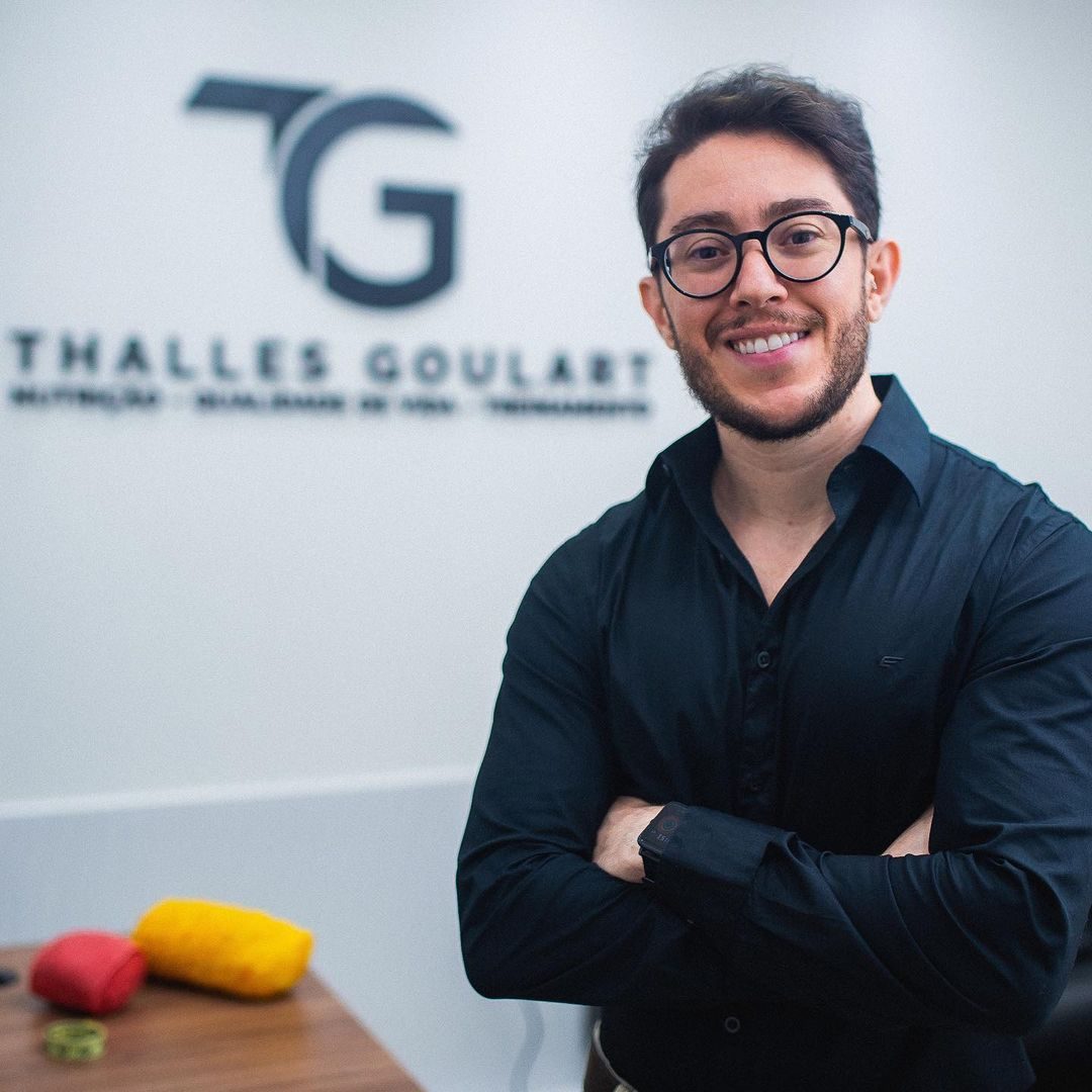 Thalles Goulart no consultório, em frente à parede com o logo TG